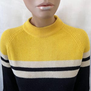 Puede incluir: Un su&eacute;ter a rayas amarillo, negro y blanco con una textura acanalada. El su&eacute;ter tiene cuello alto y mangas largas. La parte superior es amarilla, seguida de una raya blanca y negra.