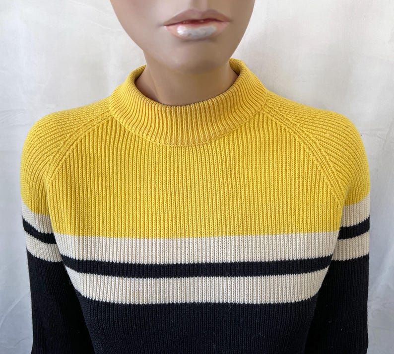 Puede incluir: Un su&eacute;ter a rayas amarillo, negro y blanco con una textura acanalada. El su&eacute;ter tiene cuello alto y mangas largas. La parte superior es amarilla, seguida de una raya blanca y negra.