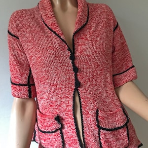 Peut inclure: Cardigan en tricot rouge et blanc avec garniture et boutons noirs. Le pull à manches courtes a deux poches avant et un col. Les bords du cardigan, des poches et des manches sont soulignés en noir.