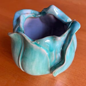 Puede incluir: Un cuenco de cerámica hecho a mano con una forma orgánica única. El cuenco presenta un esmalte turquesa brillante en el exterior y un interior azul contrastante. El diseño se asemeja a una flor o una forma plegada.