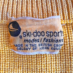 Puede incluir: Primer plano de una tela amarilla tejida con una etiqueta. La etiqueta dice "ski-doo sports modes/fashions MADE IN THE BRITISH CROW COLONY OF HONG KONG". Se ve un logotipo en blanco y negro de un esquiador.