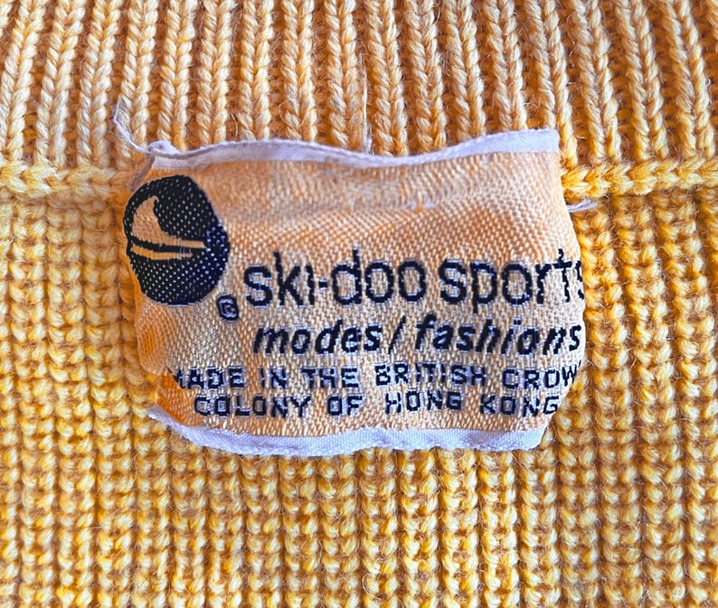 Puede incluir: Primer plano de una tela amarilla tejida con una etiqueta. La etiqueta dice "ski-doo sports modes/fashions MADE IN THE BRITISH CROW COLONY OF HONG KONG". Se ve un logotipo en blanco y negro de un esquiador.