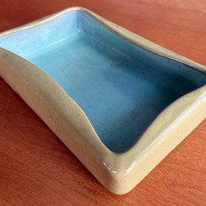 May include: A rectangular ceramic dish with a light yellow exterior and a light blue interior. The dish has a slightly curved edge on one side. The dish is empty and sits on a wooden surface.