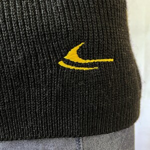 Puede incluir: Primer plano de un gorro negro de punto con un logotipo amarillo bordado en el lateral. El gorro tiene una textura acanalada y parece estar hecho de un material suave. El logotipo es un dise&ntilde;o estilizado en color amarillo brillante.