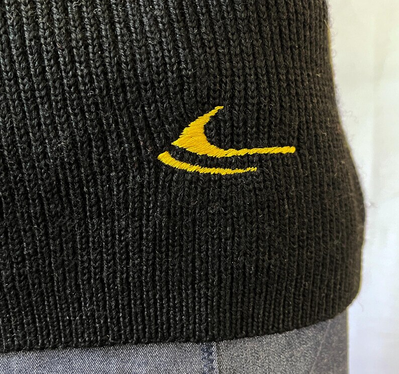 Puede incluir: Primer plano de un gorro negro de punto con un logotipo amarillo bordado en el lateral. El gorro tiene una textura acanalada y parece estar hecho de un material suave. El logotipo es un dise&ntilde;o estilizado en color amarillo brillante.