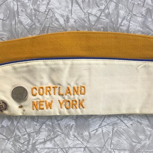 Puede incluir: Gorra de estilo militar vintage con la parte superior de color amarillo mostaza y la parte delantera de color crema. La gorra presenta texto bordado "CORTLAND NEW YORK" en hilo dorado. Un peque&ntilde;o alfiler y una moneda est&aacute;n adheridos al frente.