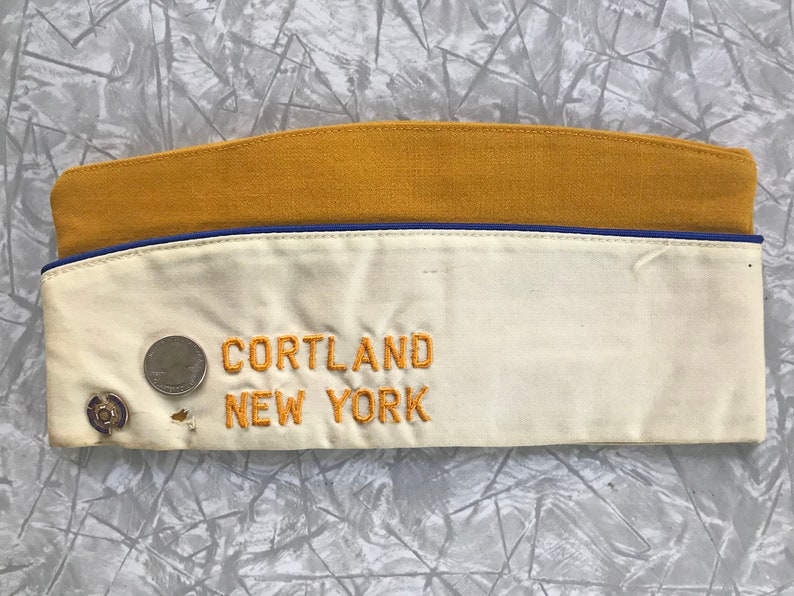 Puede incluir: Gorra de estilo militar vintage con la parte superior de color amarillo mostaza y la parte delantera de color crema. La gorra presenta texto bordado "CORTLAND NEW YORK" en hilo dorado. Un peque&ntilde;o alfiler y una moneda est&aacute;n adheridos al frente.