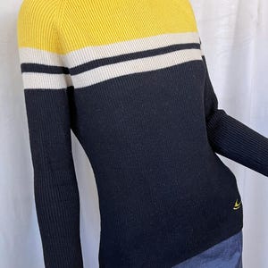 Puede incluir: Un su&eacute;ter de punto acanalado con cuello alto. La secci&oacute;n superior es amarilla, con una franja blanca, luego un cuerpo negro. Las mangas largas son negras. Un peque&ntilde;o logotipo amarillo en la parte inferior izquierda.