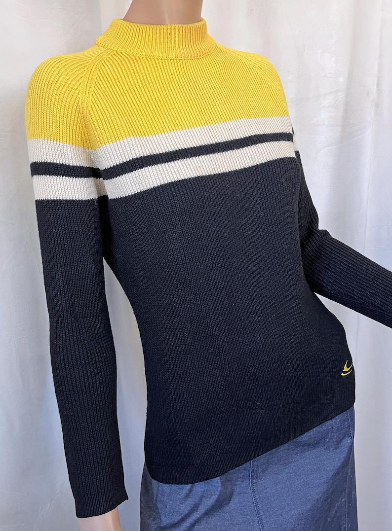 Puede incluir: Un su&eacute;ter de punto acanalado con cuello alto. La secci&oacute;n superior es amarilla, con una franja blanca, luego un cuerpo negro. Las mangas largas son negras. Un peque&ntilde;o logotipo amarillo en la parte inferior izquierda.