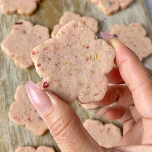 Puede incluir: Primer plano de galletas con forma de flor, de color rosa y con motas de colores. Una galleta se sostiene en una mano, y otras están dispuestas sobre una tabla de madera. Las galletas parecen ser un dulce horneado.