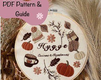 May include: An embroidery hoop featuring a fall-themed design with pumpkins, leaves, and a cozy hat. The word "Hygge" is embroidered in the center, with the phrase "Cozy & Togetherness" below. The image includes embroidery floss and wooden bobbins, and the text "PDF Pattern & Guide".