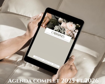 May include: A black tablet displaying a digital planner with a floral arrangement and the text "COMPLETE AGENDA 2025 AND 2026." The tablet is held by hands, resting on a cream-colored surface.