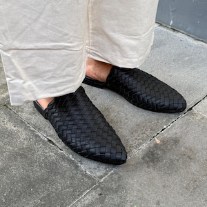 Könnte beinhalten: Schwarze gewebte Leder-Slipper. Die Schuhe haben eine spitze Zehenpartie und ein gewebtes Muster auf der Oberseite. Die Sohlen sind schwarz und die Schuhe scheinen ungetragen zu sein. Die Schuhe werden mit einer beigen Hose gezeigt.
