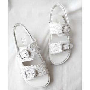 Puede incluir: Un par de sandalias de cuero blanco con un diseño tejido en las correas. Las sandalias tienen hebillas plateadas y correas ajustables. La plantilla es blanca y las sandalias están sobre un fondo blanco.