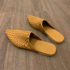 May include: A pair of woven leather tan mules with pointed toes. The shoes are made of a woven leather material and have a flat sole.
