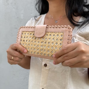 May include: A light pink leather wallet with a woven rattan center. The wallet has a flap closure and stitched edges. The design combines natural materials with a classic style, suitable for carrying cards and cash.