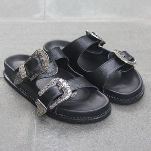 May include: A pair of black leather sandals with silver buckle accents. The sandals have two straps across the foot and a thick sole.