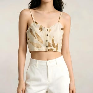 Op de afbeelding: Een beige crop top met bruine bladeren en dunne bandjes. De top heeft een knoopsluiting aan de voorkant en wordt gecombineerd met een witte broek. De stijl is casual en zomers.