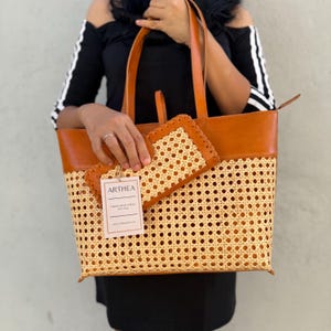 May include: A tan and brown tote bag and matching wallet. The bag has a woven rattan base and brown leather top and handles. The wallet has a similar woven design. The tag reads "ARTHEA".