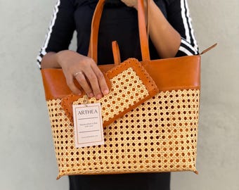 May include: A tan and brown tote bag and matching wallet. The bag has a woven rattan base and brown leather top and handles. The wallet has a similar woven design. The tag reads "ARTHEA".