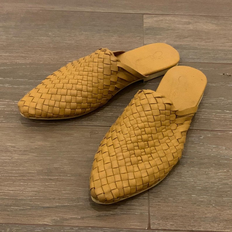 May include: A pair of woven leather mules in a light brown color. The mules have a pointed toe and a flat sole.
