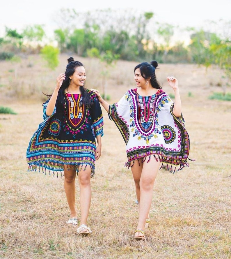 Puede incluir: Dos personas con kaftanes de patrones coloridos con flecos recorren un campo de hierba. Los kaftans tienen dise&ntilde;os geom&eacute;tricos y florales.