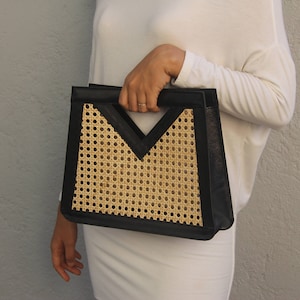 May include: Black leather handbag with a natural woven cane center panel. The bag has a geometric design with a V-shaped top and a rectangular body. The bag is held by a person wearing a white long-sleeved top.