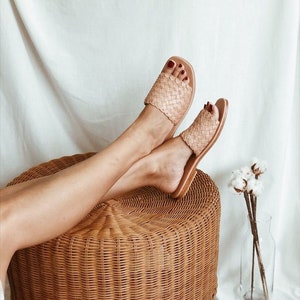 May include: A pair of woven leather sandals in a light brown color. The sandals are worn by a person sitting on a wicker stool. The sandals have a square toe and a flat sole.