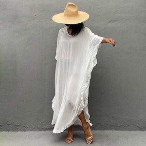 May include: A white, flowy maxi dress with a ruffled hem and short sleeves. The dress is worn with a wide-brimmed straw hat and gold sandals.