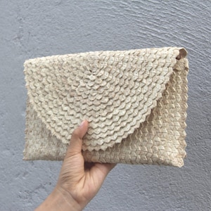 Könnte beinhalten: Eine beige gewebte Clutch mit einem gewellten Klappverschluss. Die Clutch besteht aus Naturfasern und hat ein strukturiertes Aussehen. Die Tasche wird vor einem grauen Hintergrund gehalten.