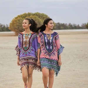 Puede incluir: Dos personas con t&uacute;nicas estampadas de colores con flecos y collares de oro. Caminan por una playa de arena.