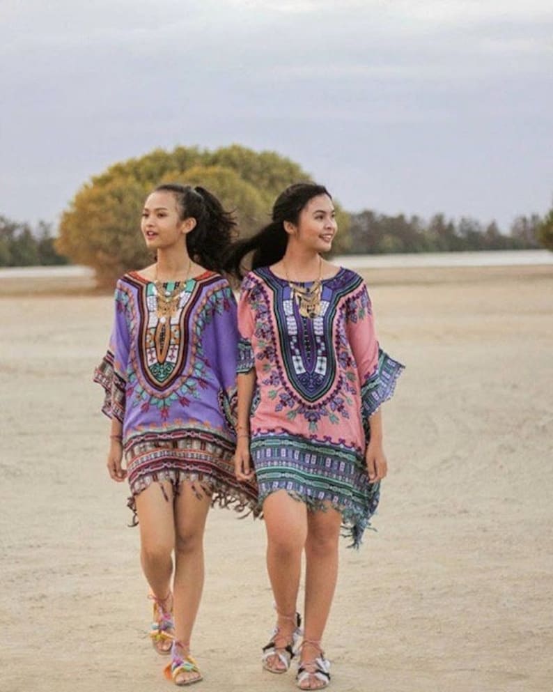 Puede incluir: Dos personas con t&uacute;nicas estampadas de colores con flecos y collares de oro. Caminan por una playa de arena.