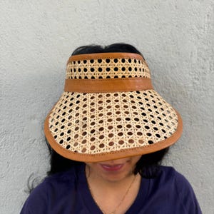May include: A brown woven visor with a leather trim. The visor is made of a woven material with a grid pattern. The visor is being worn by a person with dark hair.