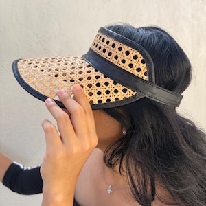 May include: A woven rattan visor with a black leather band and trim. The visor has a curved brim and a strap to secure it to the head. The rattan is a light tan colour, and the leather is black. The visor is designed to protect the eyes from the sun.