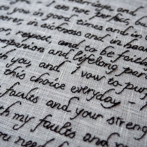 May include: Close-up of embroidered text on a light gray fabric. The black cursive script features wedding vows, including phrases like "I vow to purpose" and "in sickness and in health." The fabric has a subtle, textured appearance.