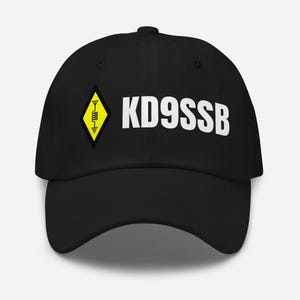 Può includere: Cappellino da baseball nero con la scritta bianca "KD9SSB" e un logo a diamante giallo con un simbolo di onda radio. Il cappellino ha una visiera curva e un bottone sulla parte superiore.