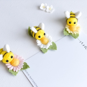 May include: Three felt bee ornaments with yellow and black stripes, each sitting on a felt flower. The flowers are white, pink, and yellow. The bees have black eyes and white wings. The background is white.