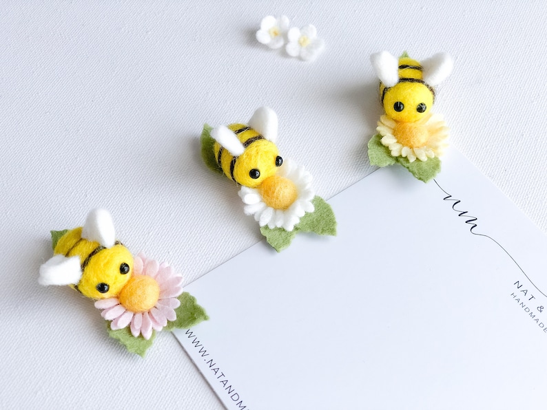 May include: Three felt bee ornaments with yellow and black stripes, each sitting on a felt flower. The flowers are white, pink, and yellow. The bees have black eyes and white wings. The background is white.