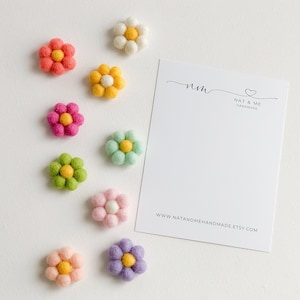 K&ouml;nnte beinhalten: Neun Filzblumen-Verzierungen in verschiedenen Farben, darunter Rosa, Gelb, Gr&uuml;n und Lila. Die Blumen haben eine gelbe Mitte und sind in einer Reihe auf wei&szlig;em Hintergrund angeordnet. Eine Visitenkarte mit dem Text "nm NAT & ME HANDMADE www.natandmehandmade.etsy.com" befindet sich in der oberen rechten Ecke des Bildes.