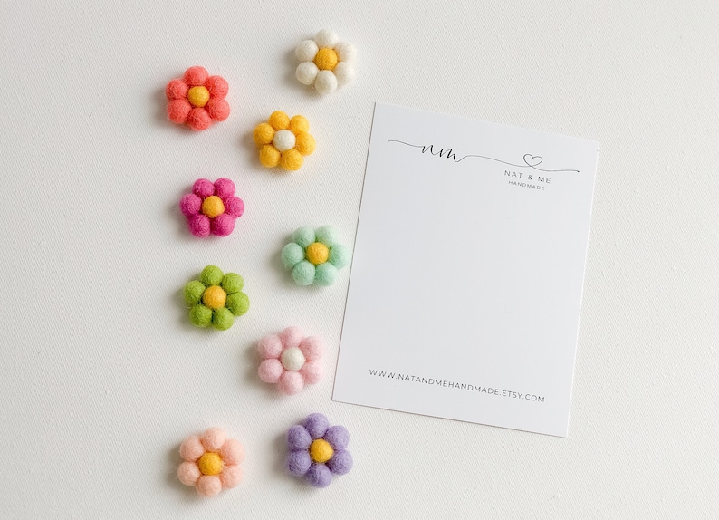 K&ouml;nnte beinhalten: Neun Filzblumen-Verzierungen in verschiedenen Farben, darunter Rosa, Gelb, Gr&uuml;n und Lila. Die Blumen haben eine gelbe Mitte und sind in einer Reihe auf wei&szlig;em Hintergrund angeordnet. Eine Visitenkarte mit dem Text "nm NAT & ME HANDMADE www.natandmehandmade.etsy.com" befindet sich in der oberen rechten Ecke des Bildes.