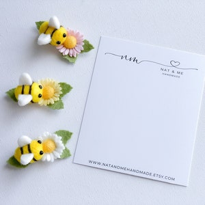 May include: Three felt bee hair clips with yellow and black stripes, white wings, and a felt flower with green leaves. The flowers are pink, yellow, and white. The card in the background says "nm Nat & Me Handmade www.natandmehandmade.etsy.com"