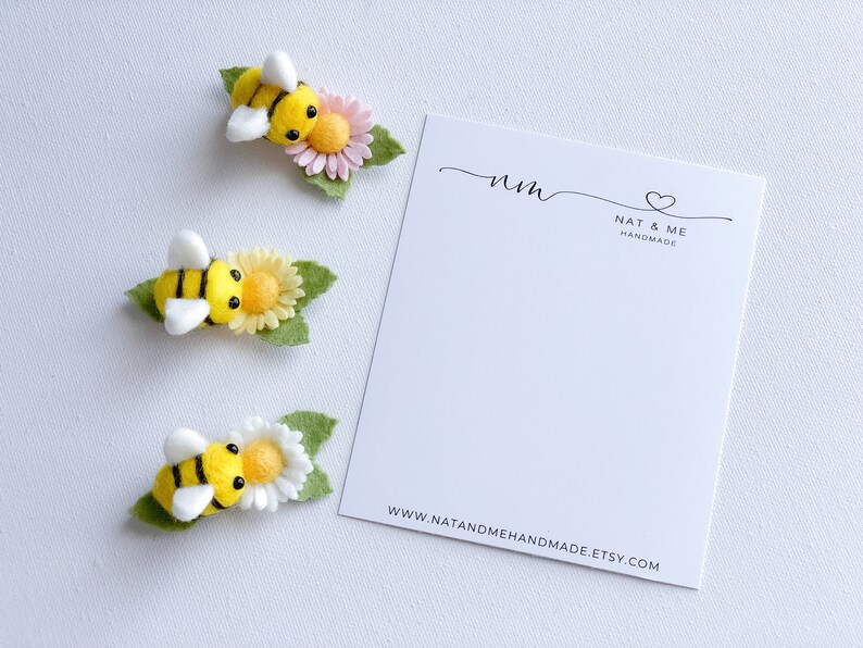 May include: Three felt bee hair clips with yellow and black stripes, white wings, and a felt flower with green leaves. The flowers are pink, yellow, and white. The card in the background says "nm Nat & Me Handmade www.natandmehandmade.etsy.com"