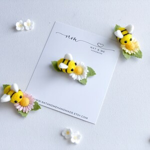 May include: Three felt bee brooches with yellow and black stripes, white wings, and black eyes. Each bee is sitting on a felt flower with green leaves. The flowers are white with yellow centers. The brooches are displayed on a white card with the text "Nat & Me Handmade" and a website address.