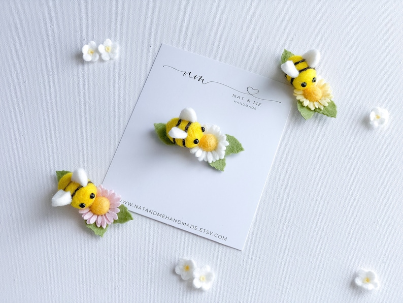 May include: Three felt bee brooches with yellow and black stripes, white wings, and black eyes. Each bee is sitting on a felt flower with green leaves. The flowers are white with yellow centers. The brooches are displayed on a white card with the text "Nat & Me Handmade" and a website address.