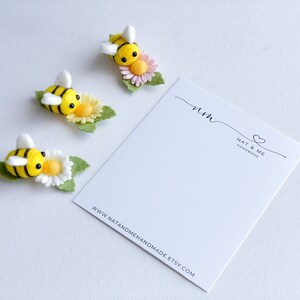 May include: Three felt bumblebees with yellow and black stripes are perched on white and yellow flowers with green leaves. The bees have black eyes and white wings. The flowers have pink centers. The bees are arranged in a row on a white background. The image also includes a white card with the text "Nat & Me Handmade" and a website address.