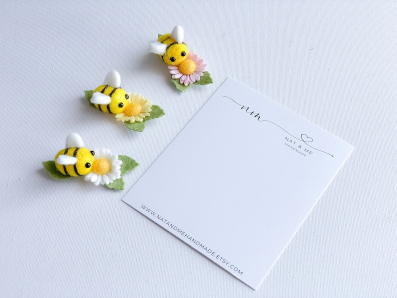 May include: Three felt bumblebees with yellow and black stripes are perched on white and yellow flowers with green leaves. The bees have black eyes and white wings. The flowers have pink centers. The bees are arranged in a row on a white background. The image also includes a white card with the text "Nat & Me Handmade" and a website address.
