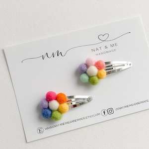 K&ouml;nnte beinhalten: Zwei Regenbogen-Pompon-Haarklammern auf einer wei&szlig;en Karte mit dem Text "nm NAT & ME HANDMADE" und Website-Informationen.