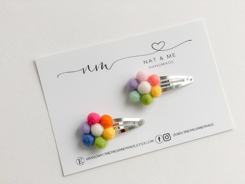 K&ouml;nnte beinhalten: Zwei Regenbogen-Pompon-Haarklammern auf einer wei&szlig;en Karte mit dem Text "nm NAT & ME HANDMADE" und Website-Informationen.