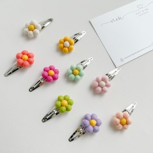 K&ouml;nnte beinhalten: Sortiment aus zehn Filzblumen-Haarklammern in verschiedenen Farben, darunter Wei&szlig;, Gelb, Rosa, Gr&uuml;n und Lila. Jede Blume hat eine kleine gelbe Mitte. Die Klammern sind silberfarben und haben ein schlichtes Design.