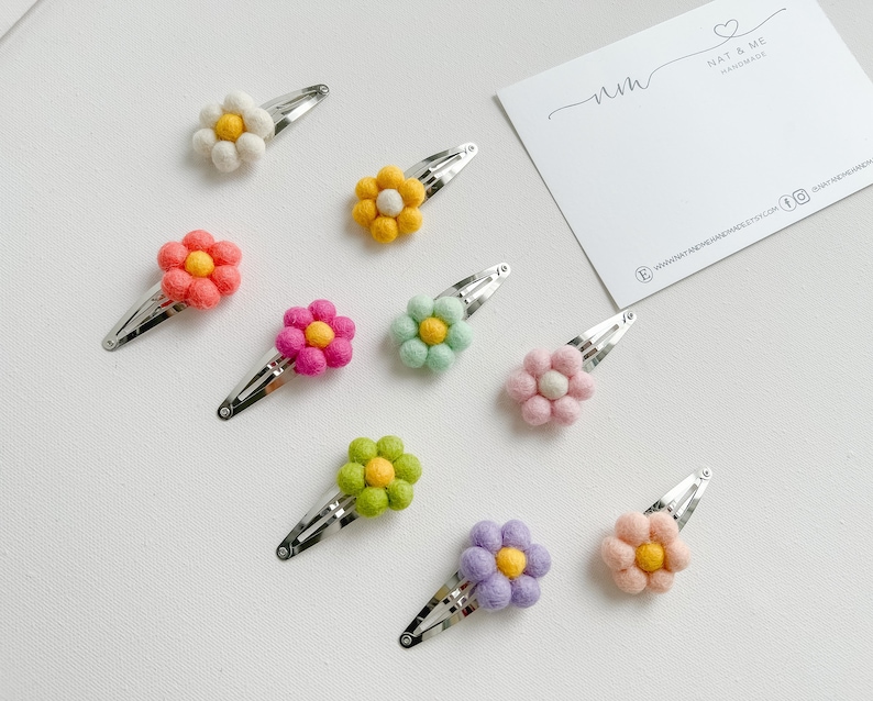 K&ouml;nnte beinhalten: Sortiment aus zehn Filzblumen-Haarklammern in verschiedenen Farben, darunter Wei&szlig;, Gelb, Rosa, Gr&uuml;n und Lila. Jede Blume hat eine kleine gelbe Mitte. Die Klammern sind silberfarben und haben ein schlichtes Design.